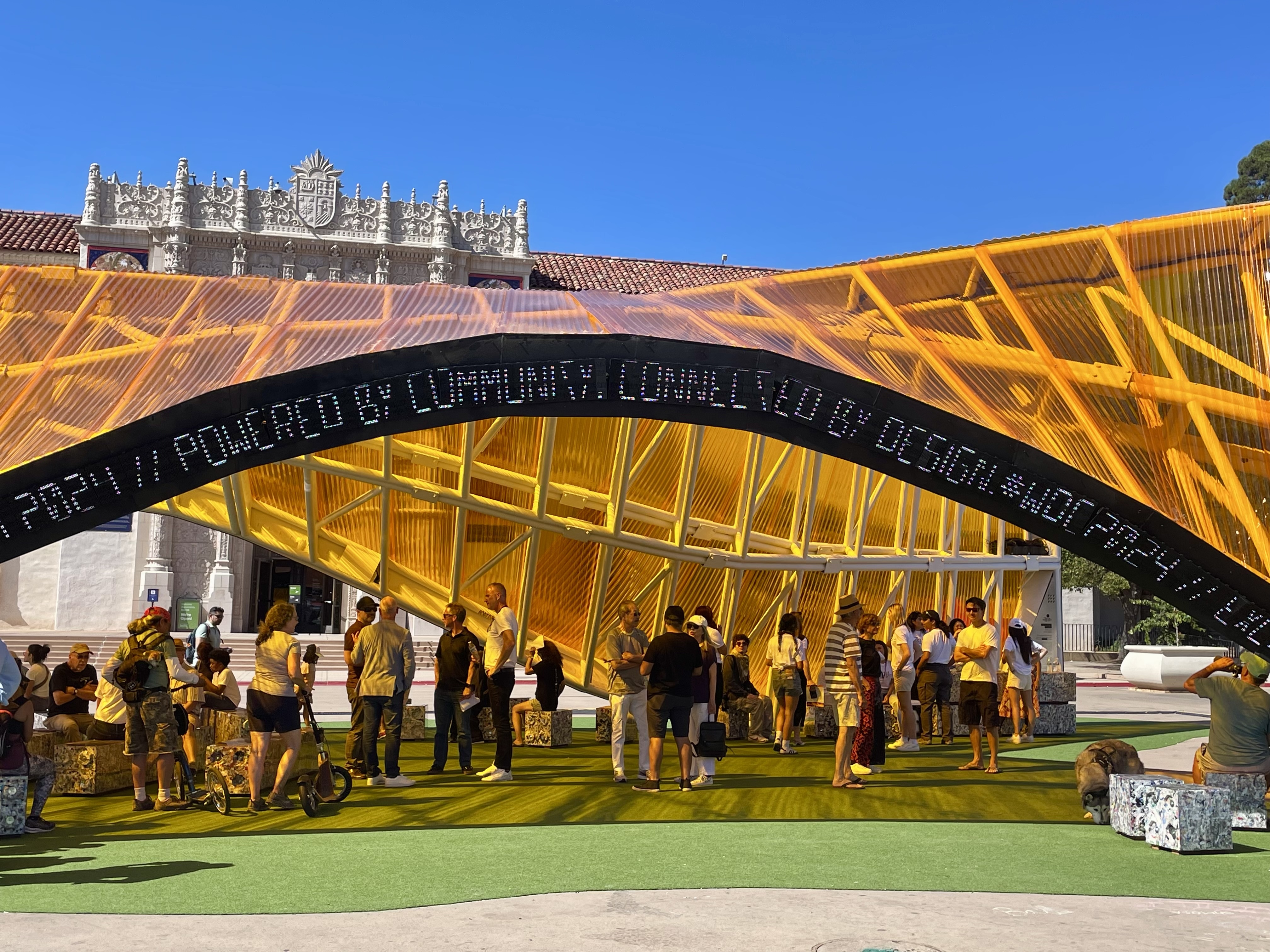 Poetry Exchange Pavilion at Balboa Park, San Diego — a large arched structure with yellow lattice roof and a scrolling LED text band, surrounded by visitors.